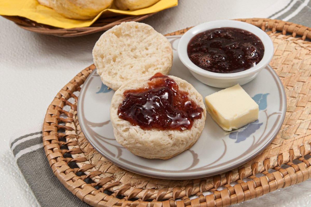 This photo of the biscuits show one cut open with jam on it on a wicker placemat with a basket of biscuits in the background.