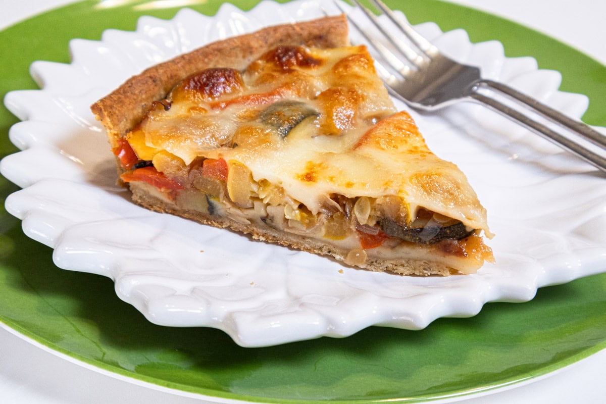This picture of the Summer Vegetable Tart shows a slice of the tart on a white daisy plate on a green background with a fork.
