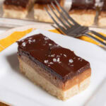 A Millionaires Shortbread bar sits on a white plate with a fork and a tray of bars behind it.