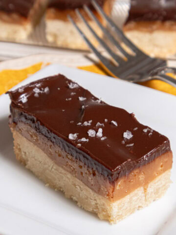 A Millionaires Shortbread bar sits on a white plate with a fork and a tray of bars behind it.