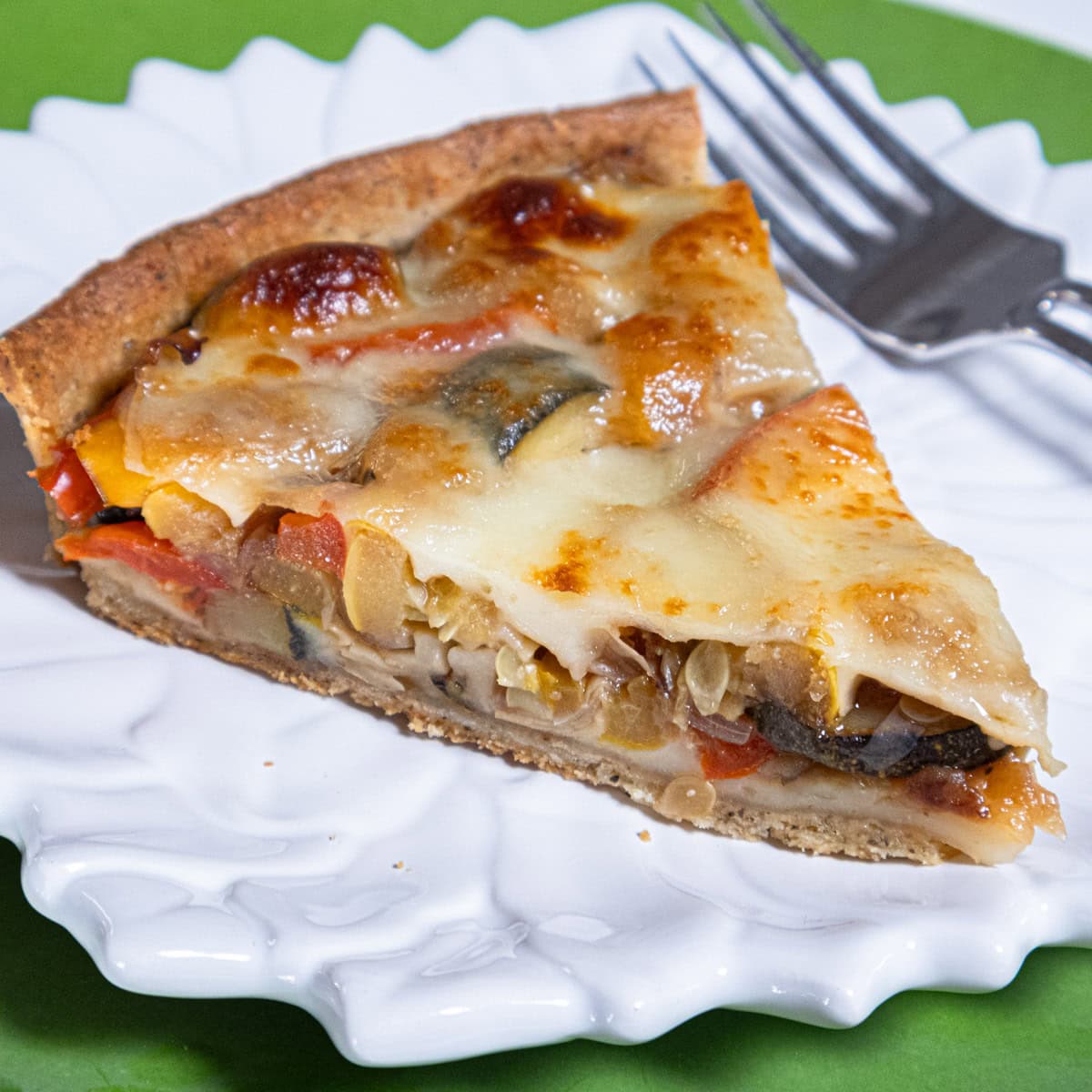 A slice of the Summer Vegetable Tart sits on a white plate with a fork on a green background.