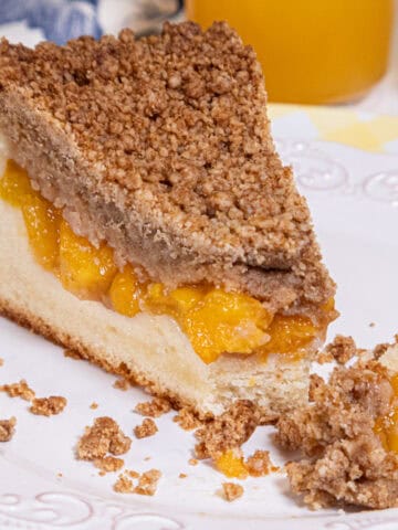 A slice of Peach Crumb Coffee Cake sits on a white plate and yellow plate with a blue and white napkin and a glass of peach juice in the background.