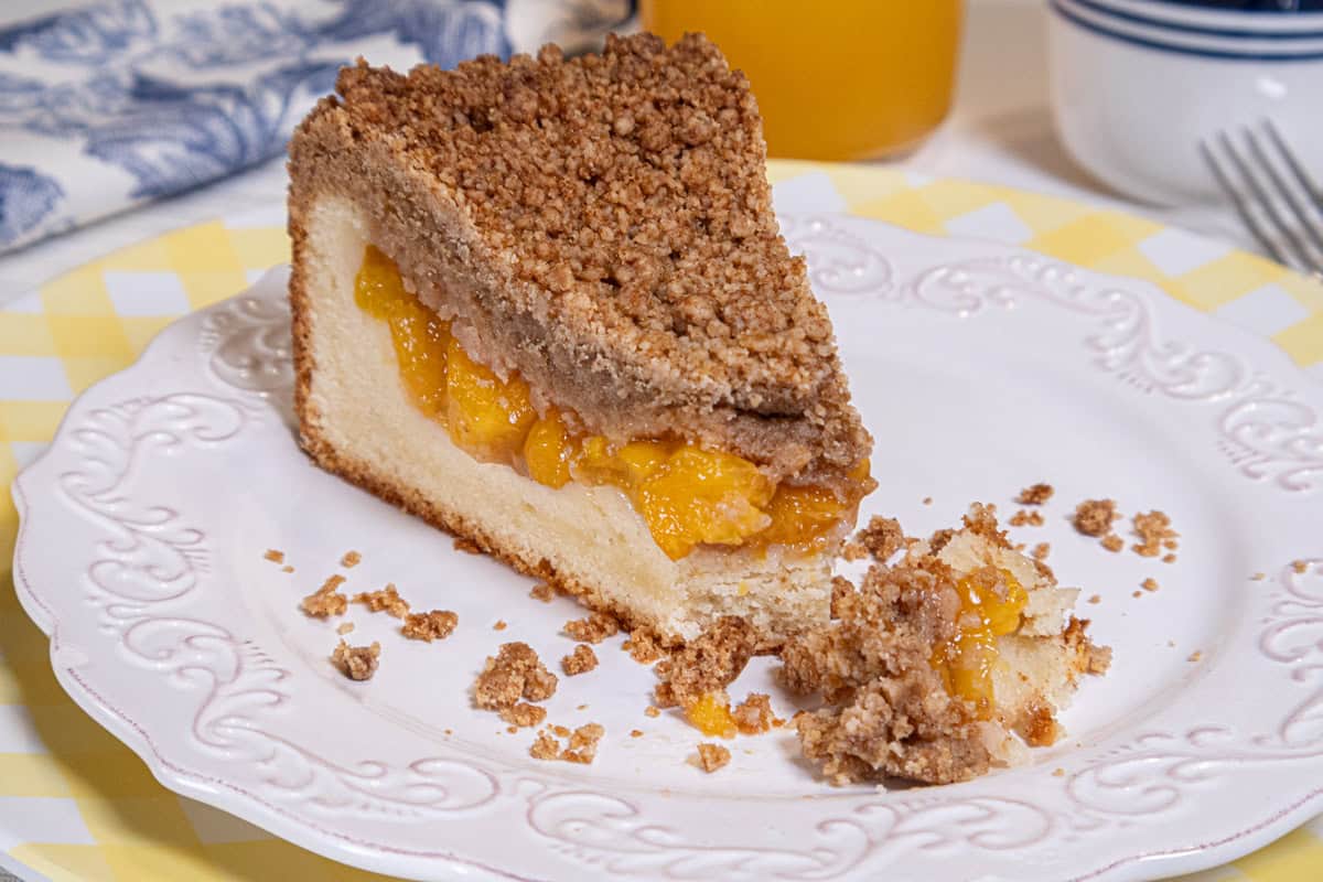 A slice of Peach Crumb Coffee Cake sits on a white plate and yellow plate with a blue and white napkin and a glass of peach juice in the background.