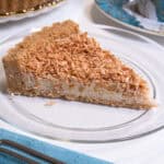 A slice of Tropical Macadamia & Coconut Tart sits on a clear plate with a teal colored napkin and cup and saucer beside it.