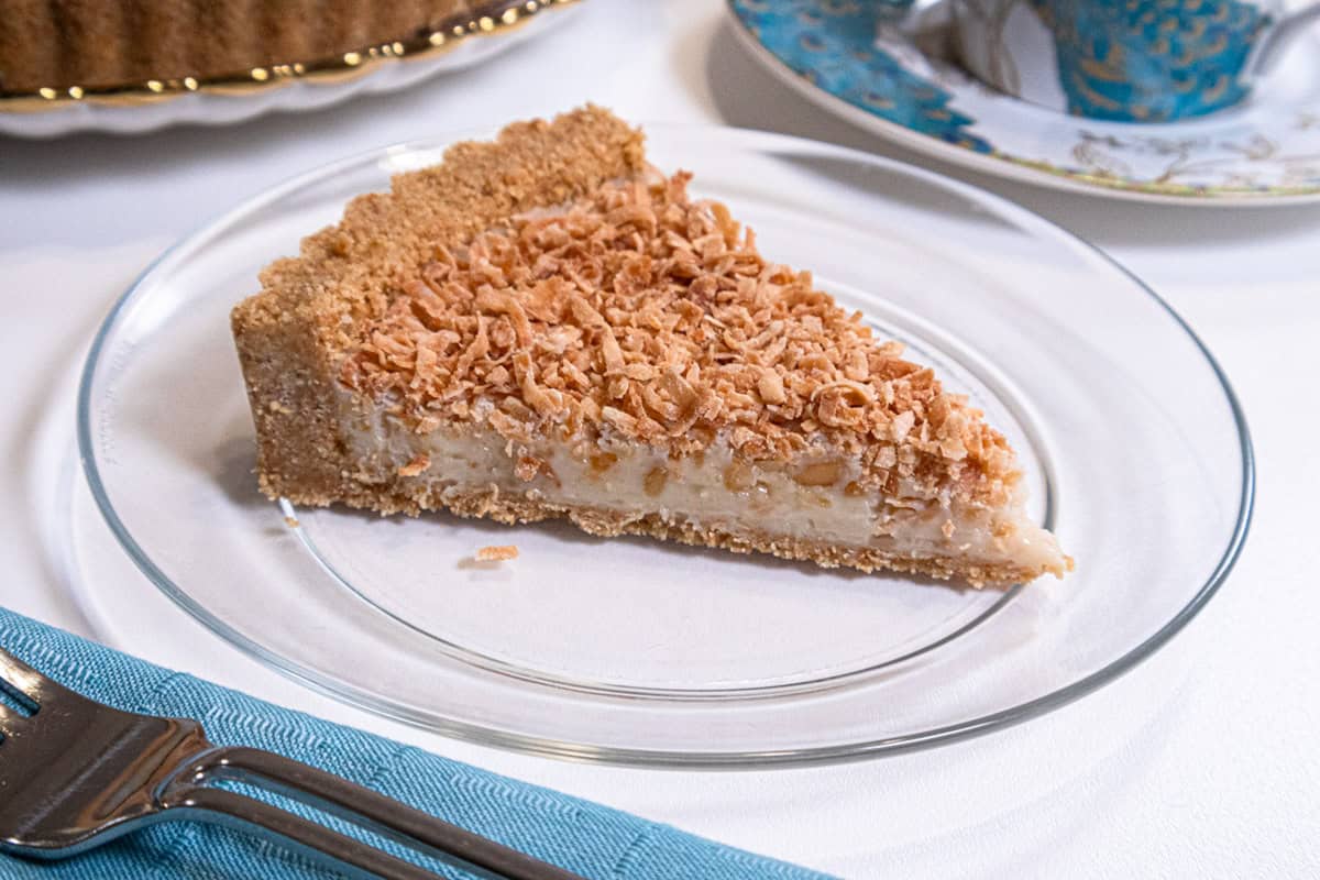 A slice of Tropical Macadamia & Coconut Tart sits on a clear plate with a teal colored napkin and cup and saucer beside it.
