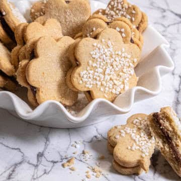 A white bowl holds single Spiced Shortbread cookies on a marble background with a broken sandwiched cookie in the foreground.