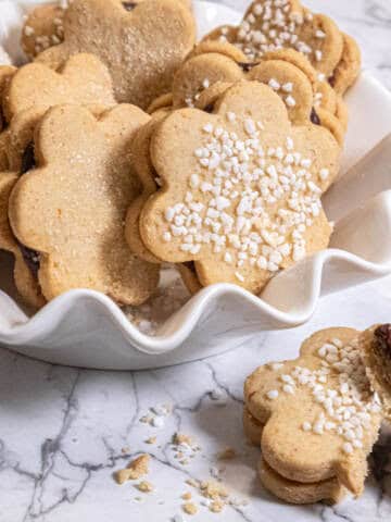 A white bowl holds single Spiced Shortbread cookies on a marble background with a broken sandwiched cookie in the foreground.