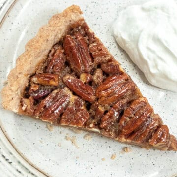 A slice of the Butter Run Pecan Tart sists on a brown striped plate with a dollup of whipped cream.