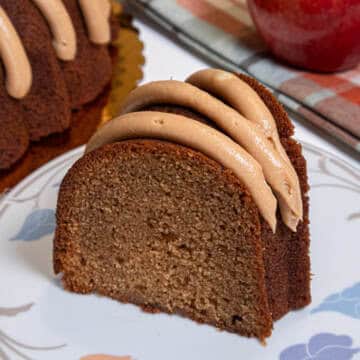 A slice of the Caramel Apple Spiced Cake sits on a white plate with colored leaves with an apple, plaid napkin and the cake in the background.