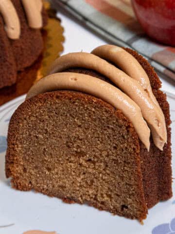 A slice of the Caramel Apple Spiced Cake sits on a white plate with colored leaves with an apple, plaid napkin and the cake in the background.