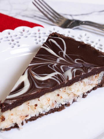A slice of the Chocolate Coconut Tart sits on a lace edged white plate with a red napkin and a fork behind it.