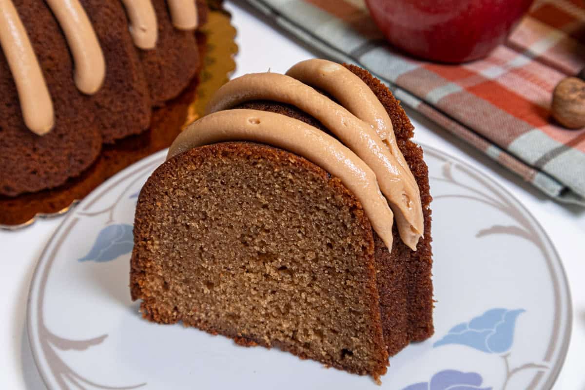 A slice of the Caramel Apple Spiced Cake sits on a white plate with colored leaves with an apple, plaid napkin and the cake in the background.