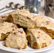Eight Pumpkin Pecan Scones sit on a white plate with mugs and spoons in the background.