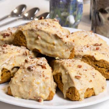 Eight Pumpkin Pecan Scones sit on a white plate with mugs and spoons in the background.