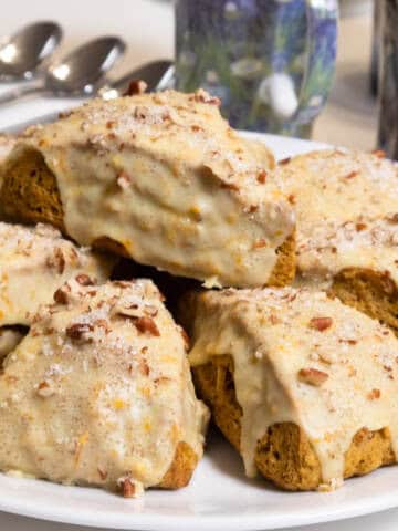 Eight Pumpkin Pecan Scones sit on a white plate with mugs and spoons in the background.