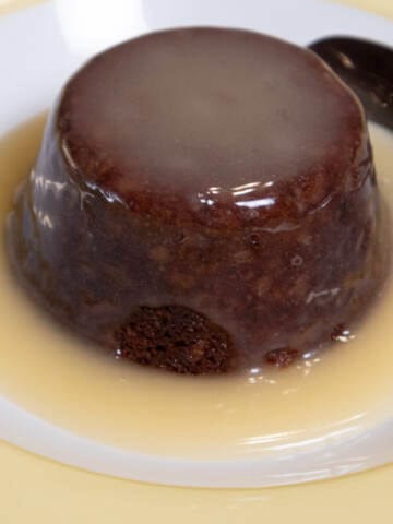 An individual Sticky Toffee Pudding sits in a puddle of Lemon Sauce in a yellow and white bowl.