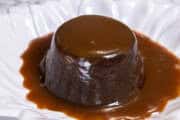 This photograph shows a sticky toffee pudding enveloped in toffee sauce on a white plate.