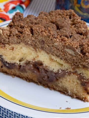 A slice of the Apple Cinnamon Coffee Cake sits on a yellow rimmed plate with a napkin in the back ground.