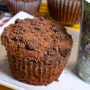 A whole coffee cake muffin sits on a wite plate with a colored mug on the plate and more muffins in the background.