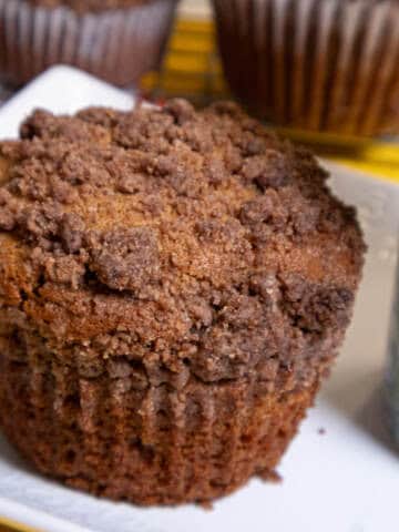 A whole coffee cake muffin sits on a wite plate with a colored mug on the plate and more muffins in the background.