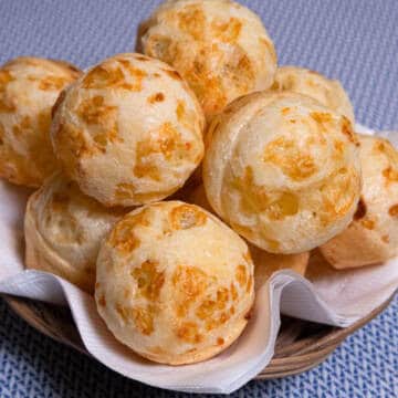 Balls of Brazilian Cheese Bread sit in a wicker basket in a white napkiin on a blue background.