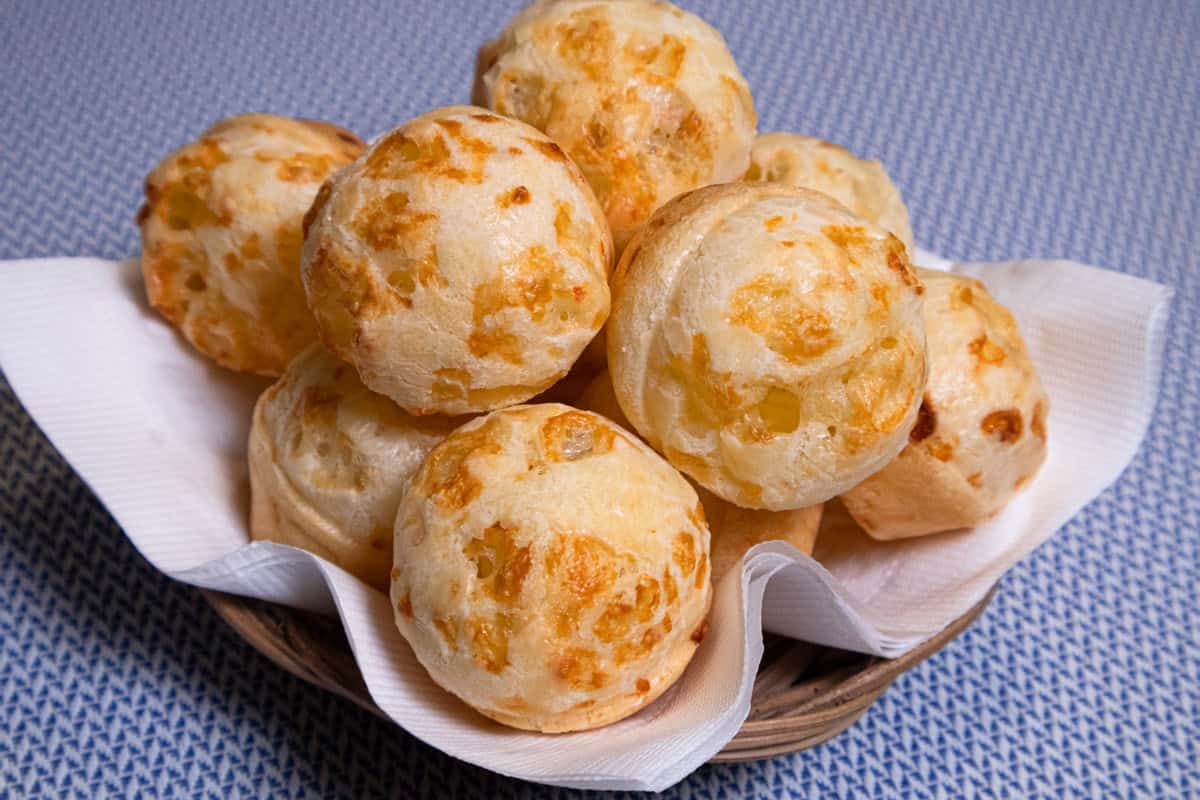 Balls of Brazilian Cheese Bread sit in a wicker basket in a white napkiin on a blue background.