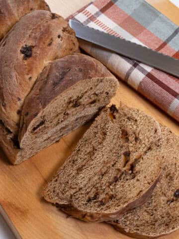 Slices of the Rye Raisin Bread sits on a cutting board with the whole behind it surrounded by a plaid towel and knife.