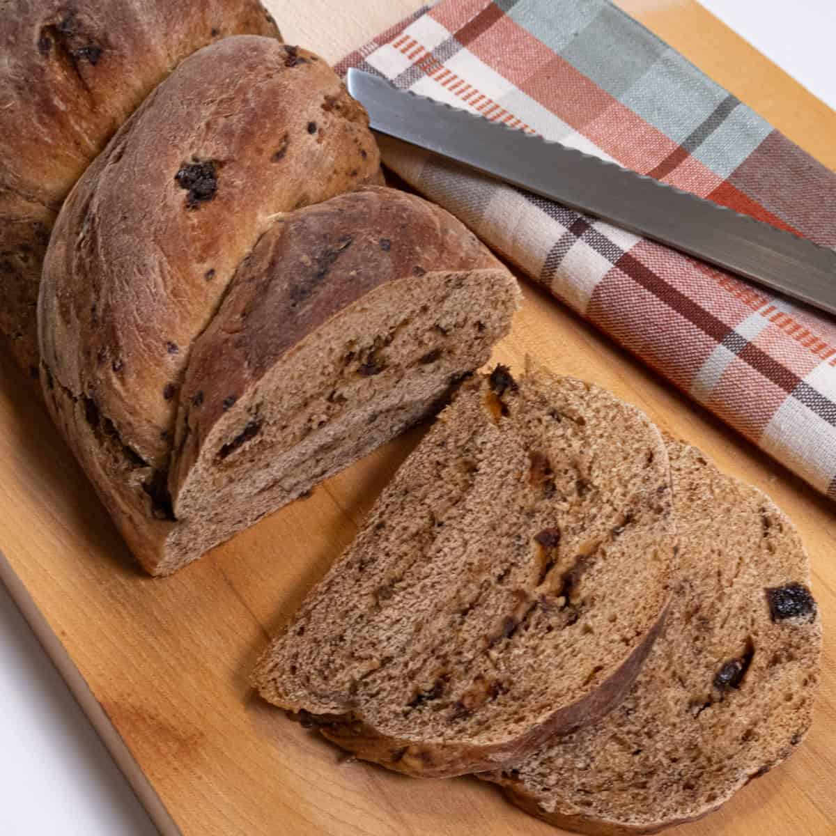 Slices of the Rye Raisin Bread sits on a cutting board with the whole behind it surrounded by a plaid towel and knife. 