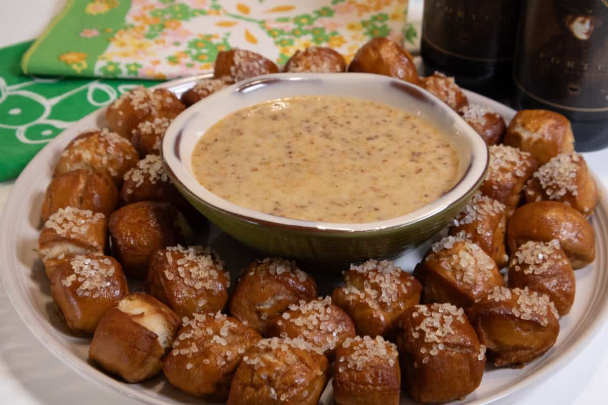 Pretzel bites surround a bowl of Hot Honey Mustard dip with napkins in the background on bottles of beer on the side.