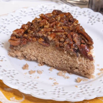 A slice of the pecan upside down cake sits on a white plate with a lace edge which sits o ntop of another orangish pate.