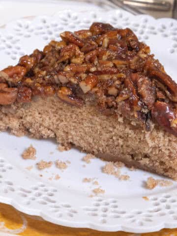 A slice of the pecan upside down cake sits on a white plate with a lace edge which sits o ntop of another orangish pate.