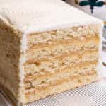 The Lemon Layer Cake is on a clear plate with a vase in the background and a serving utensil.