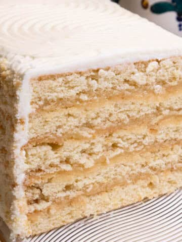 The Lemon Layer Cake is on a clear plate with a vase in the background and a serving utensil.