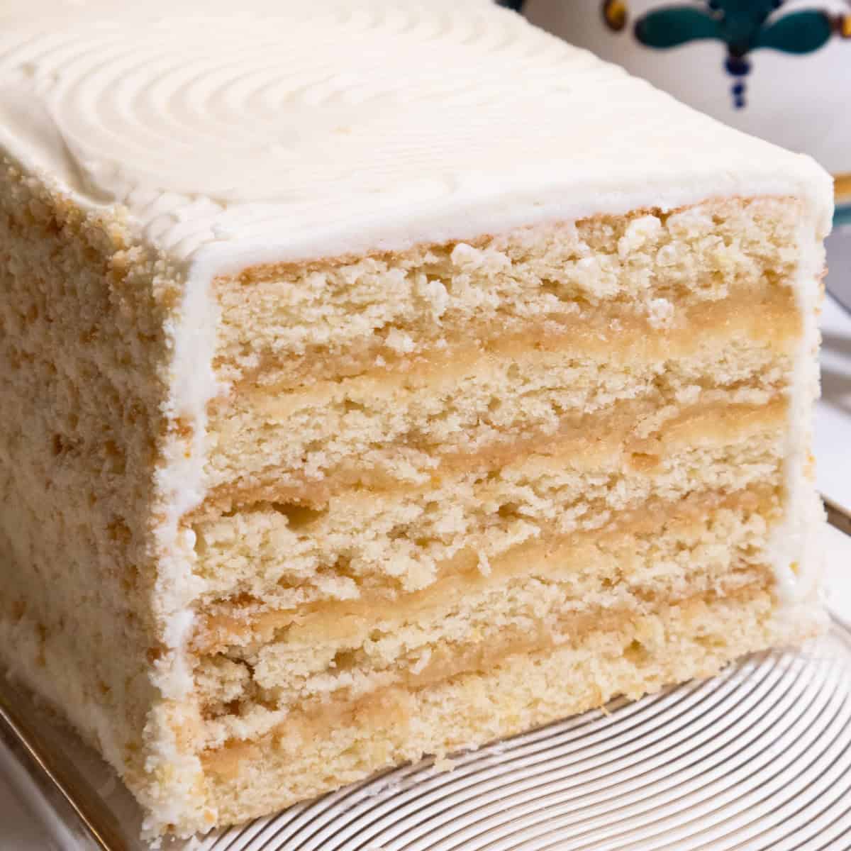 The Lemon Layer Cake is on a clear plate with a vase in the background and a serving utensil. 