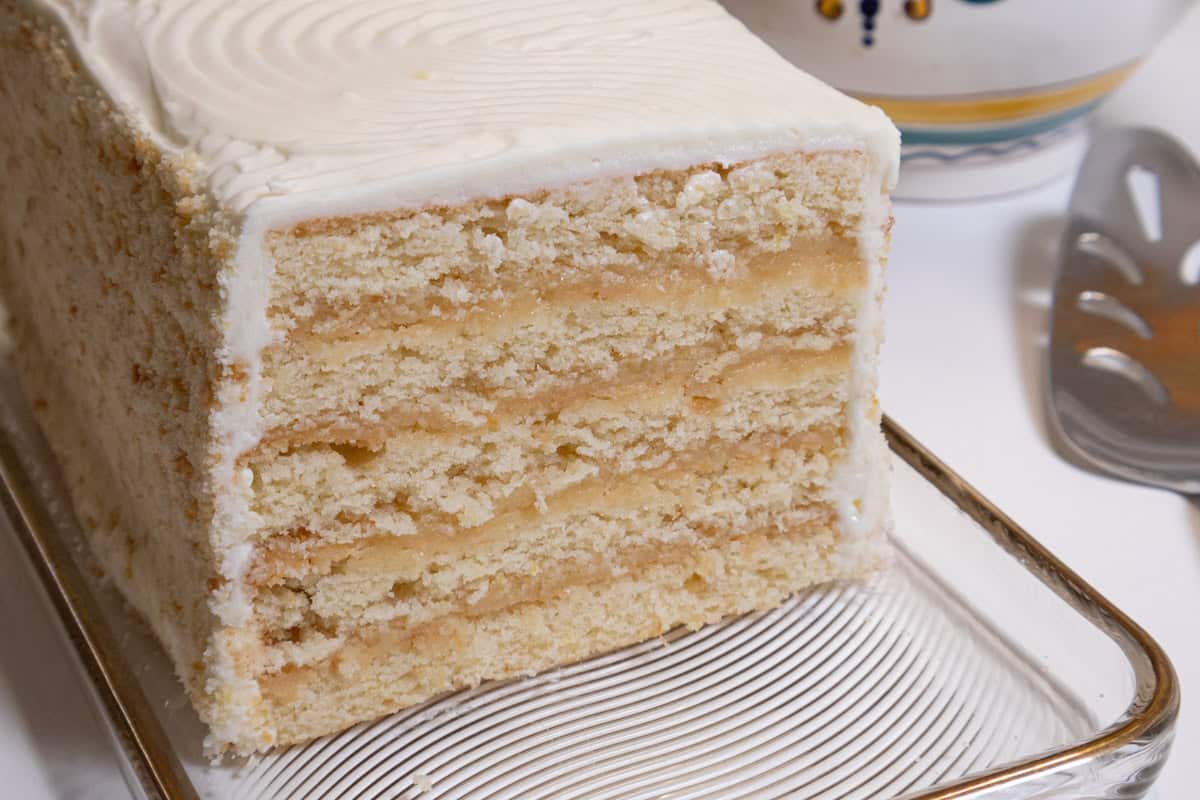 The Lemon Layer Cake is on a clear plate with a vase in the background and a serving utensil.