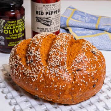 This orange hued bread sits on a white lattice plate with a bottle of calamata olives and roasted red pepper is in the background.