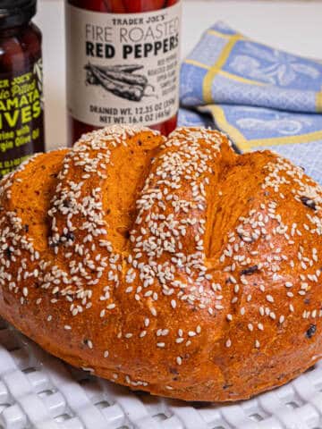 This orange hued bread sits on a white lattice plate with a bottle of calamata olives and roasted red pepper is in the background.