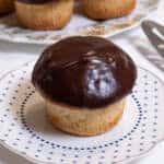 A Boston Cream Cupcake sits o a small blue and white plate with a server on the side and more cupcakes in the background.