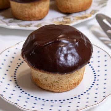 A Boston Cream Cupcake sits o a small blue and white plate with a server on the side and more cupcakes in the background.