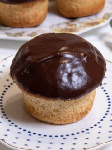 A Boston Cream Cupcake sits o a small blue and white plate with a server on the side and more cupcakes in the background.