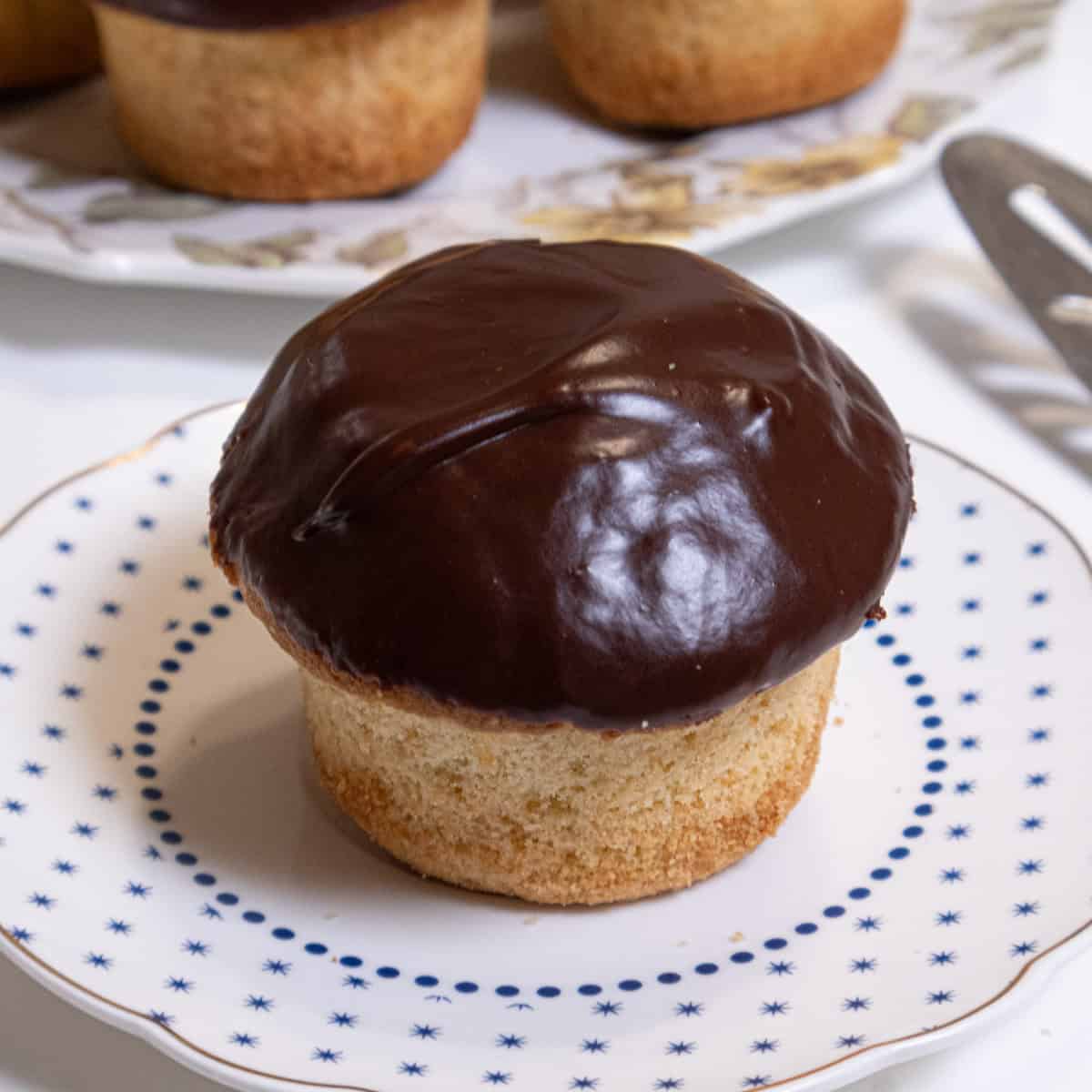 A Boston Cream Cupcake sits o a small blue and white plate with a server on the side and more cupcakes in the background.