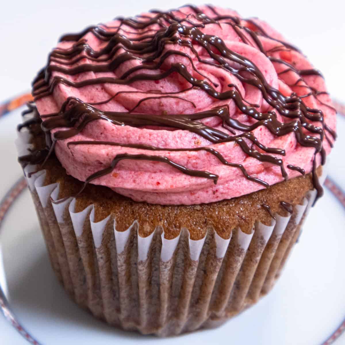A Banana Split Cupcake with its strawberry buttercream drizzled with chooolate sits on a white plate .