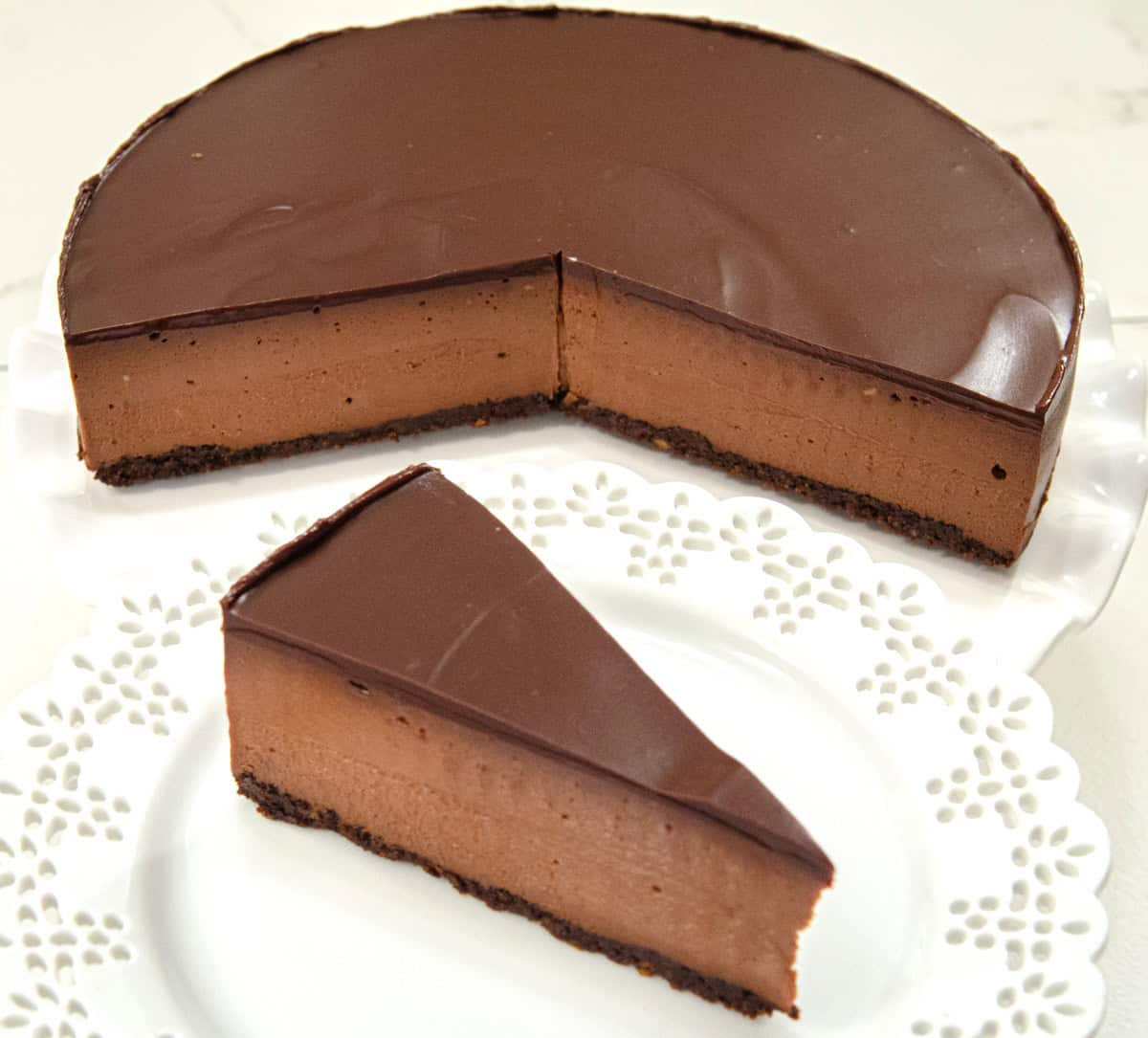 A slice of the Triple Chocolate Cheesecake sits on a lace edged white plate with the cut cheesecake in the background.