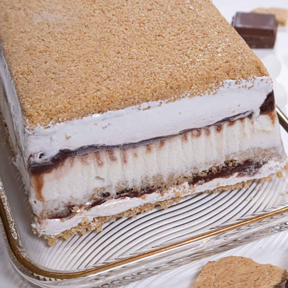 The S'Mores Ice Cream Cake sits on a gold rimmed cake plate with shards of graham cracker crums and chocolate around it, 