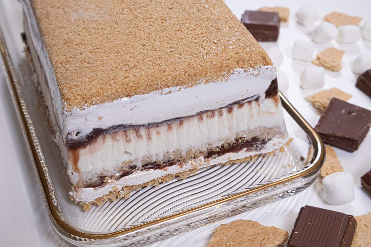 The S'Mores Ice Cream Cake sits on a gold rimmed cake plate with shards of graham cracker crumbs and chocolate around it,
