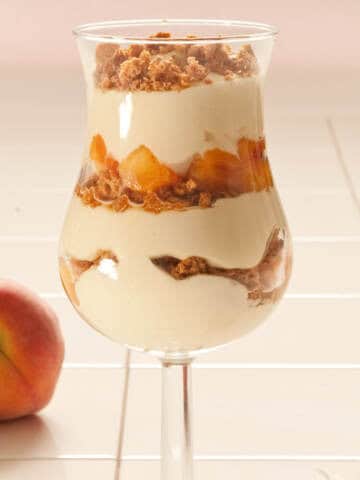 A parfait glass is filled with mascarpone filling, amaretti cookies, and fresh peaches with a fresh peach i the backgrond.