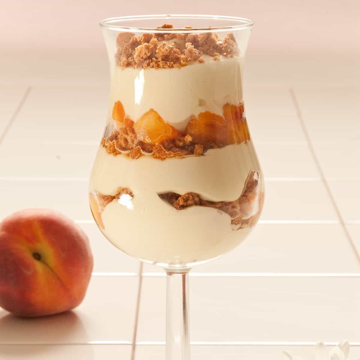 A parfait glass is filled with mascarpone filling, amaretti cookies, and fresh peaches with a fresh peach i the backgrond.