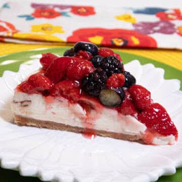 A slice of the No Bake White Chocolate Cheesecake with a mixed berry topping sets on a white plate with a green charger and color flowered napkin.