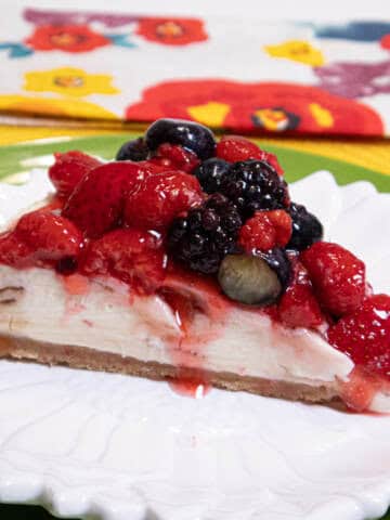 A slice of the No Bake White Chocolate Cheesecake with a mixed berry topping sets on a white plate with a green charger and color flowered napkin.