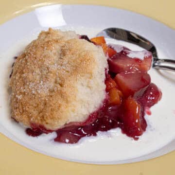 This photo for the Peach and Plum Cobbler shows the fruit in a bowl with a biscuit on top swimming in cream.
